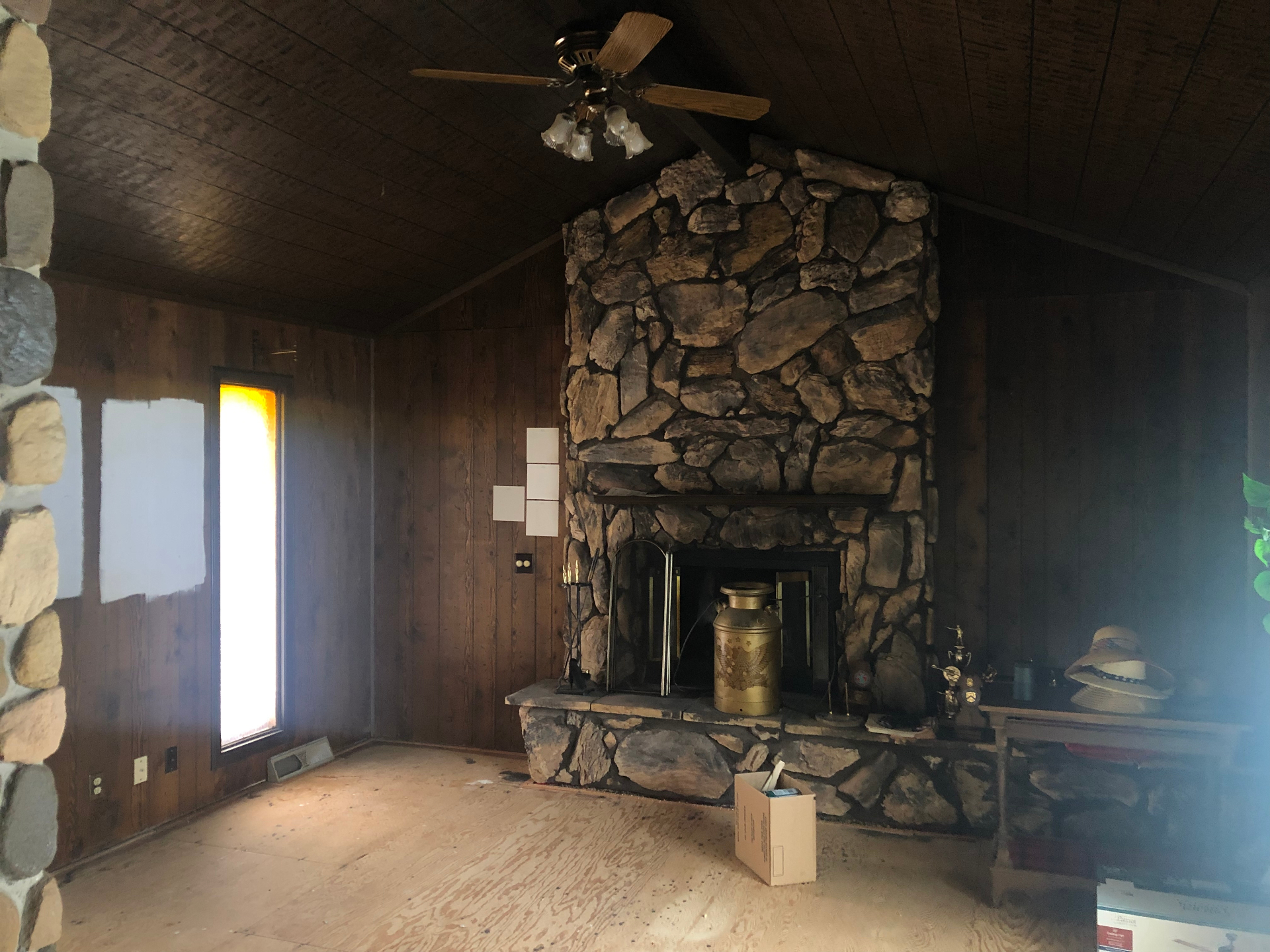 Cleared living room with dark wood plank ceiling, dark wood paneling, and a floor-to-ceiling stone fireplace, with paint test squares on the wall
