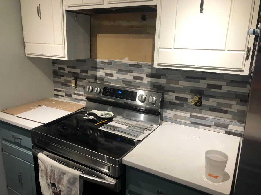 Kitchen with a newly installed grey, black, and white linear tile backsplash behind a range and white upper cabinets