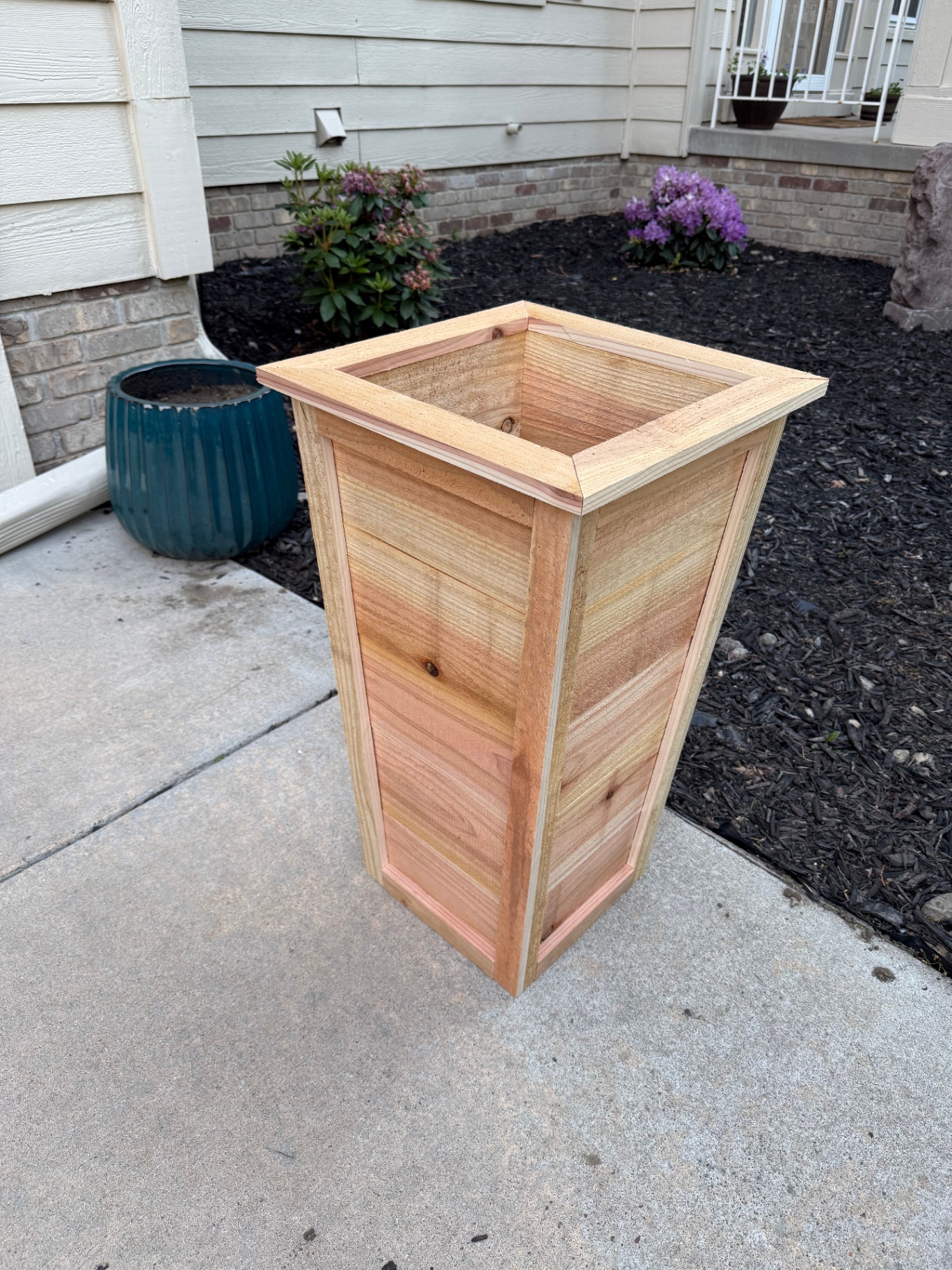 Tall tapered cedar planter box with mitered trim and horizontal slat panels sitting on a front walkway