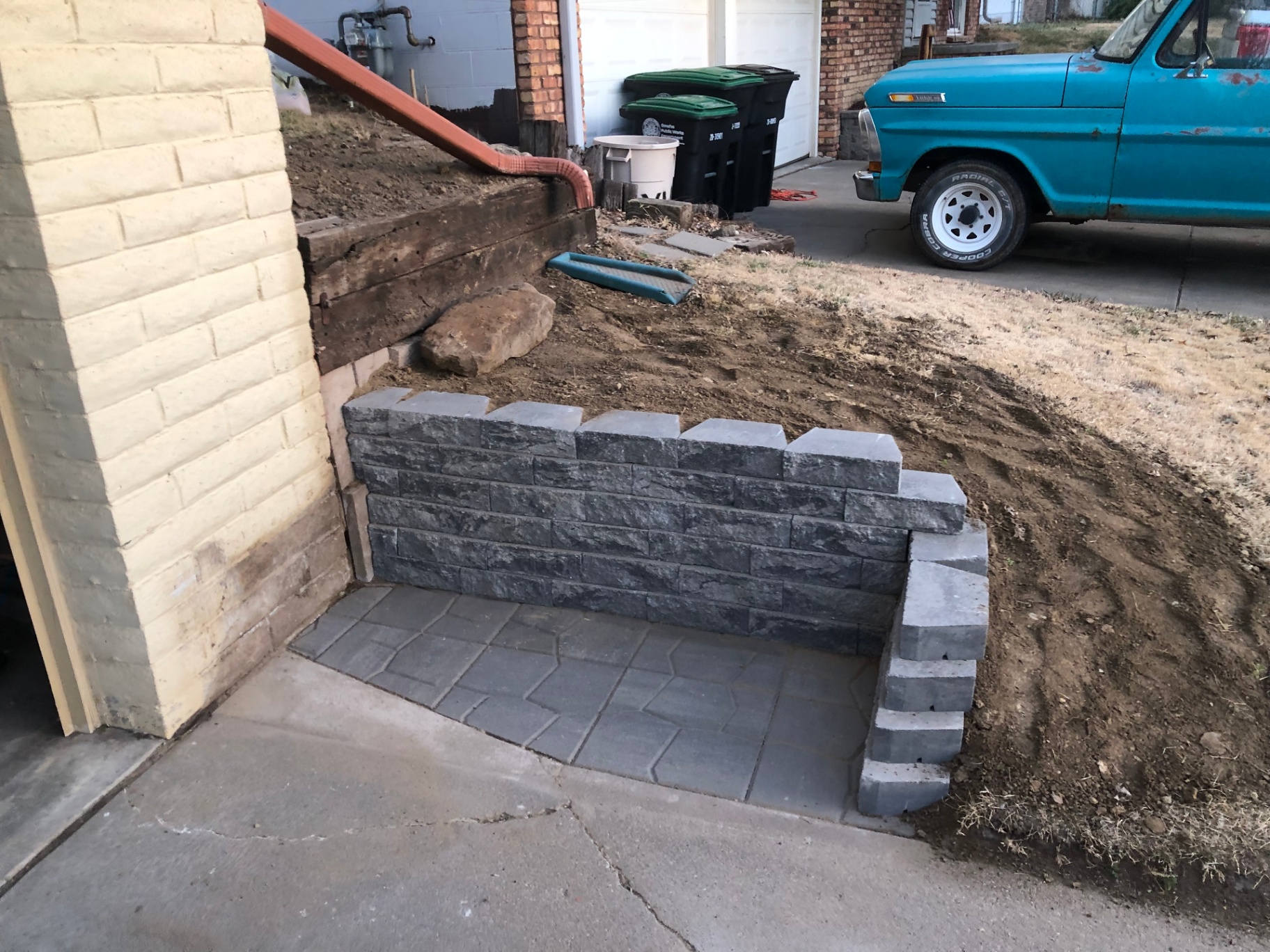 Finished stacked-block retaining wall and paver pad tucked against a painted brick garage wall