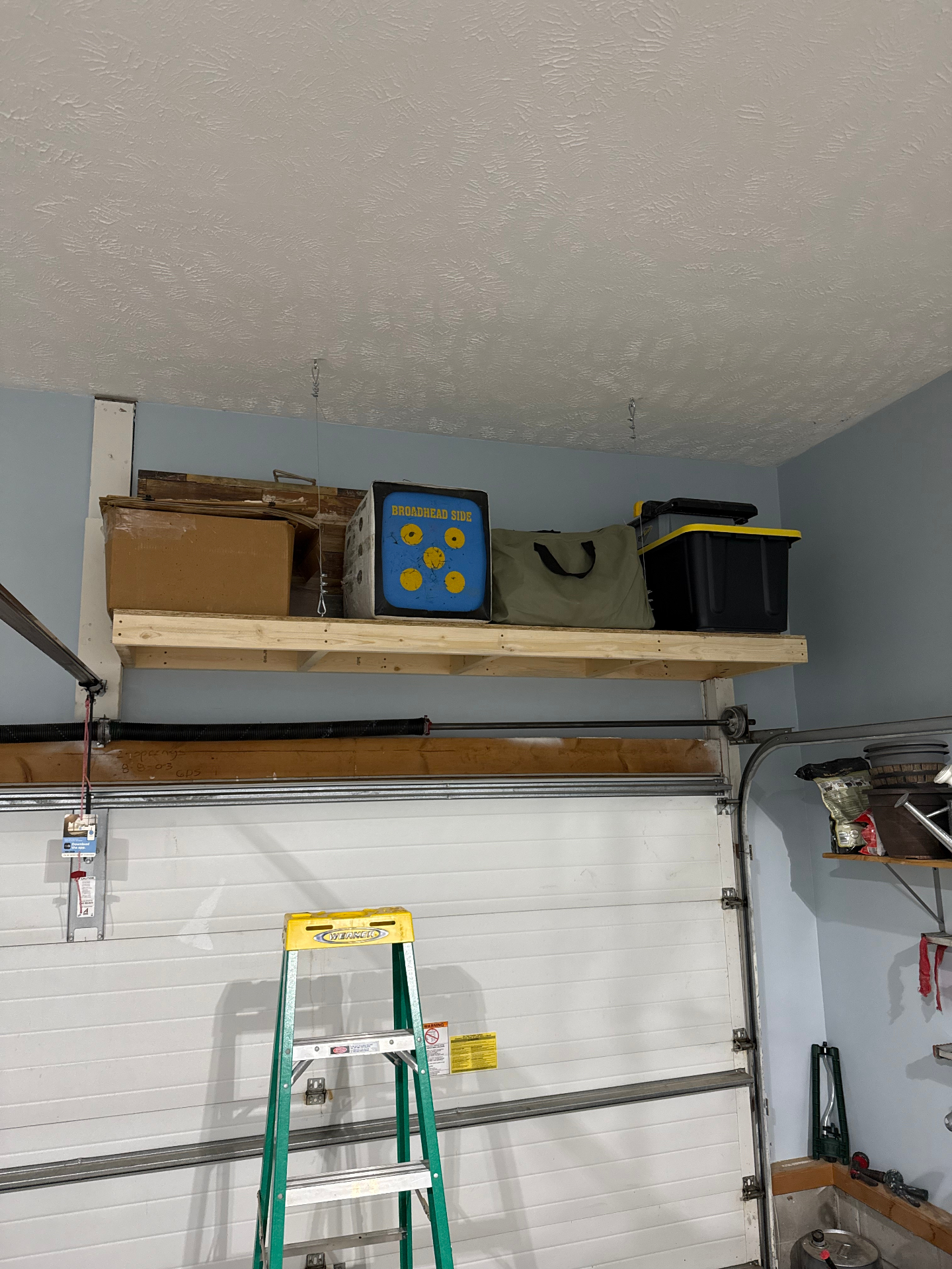 Overhead garage storage shelf holding boxes, a target block, a duffel bag, and a black tote above the garage door