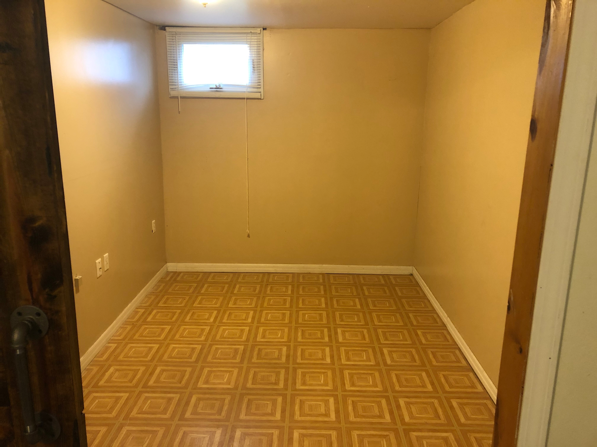 Empty basement room with yellowed walls and dated square-pattern parquet-look vinyl flooring, before renovation