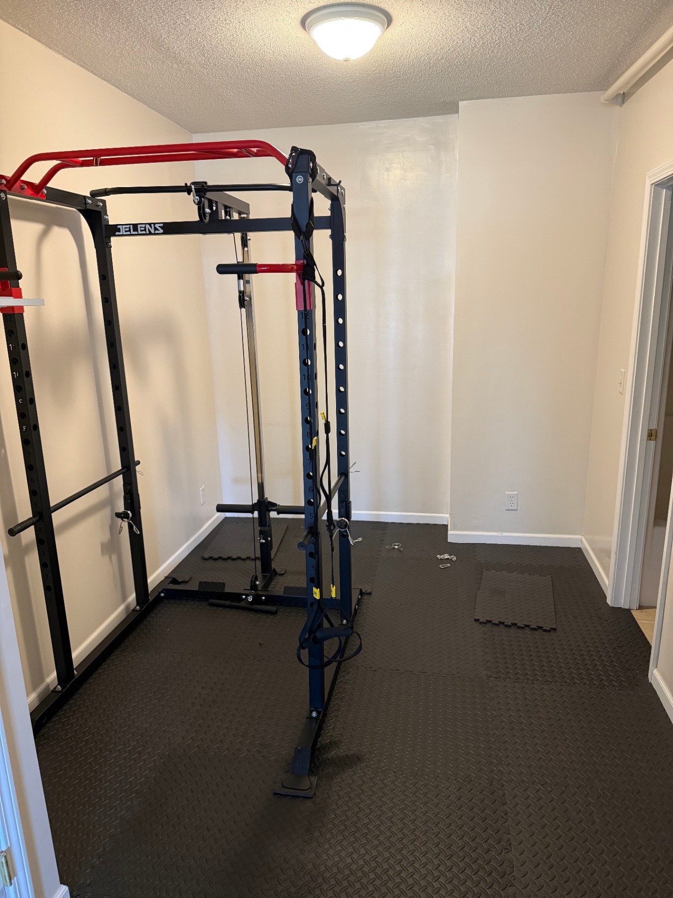 Small workout room with plain white walls and a power rack sitting on interlocking rubber floor tiles