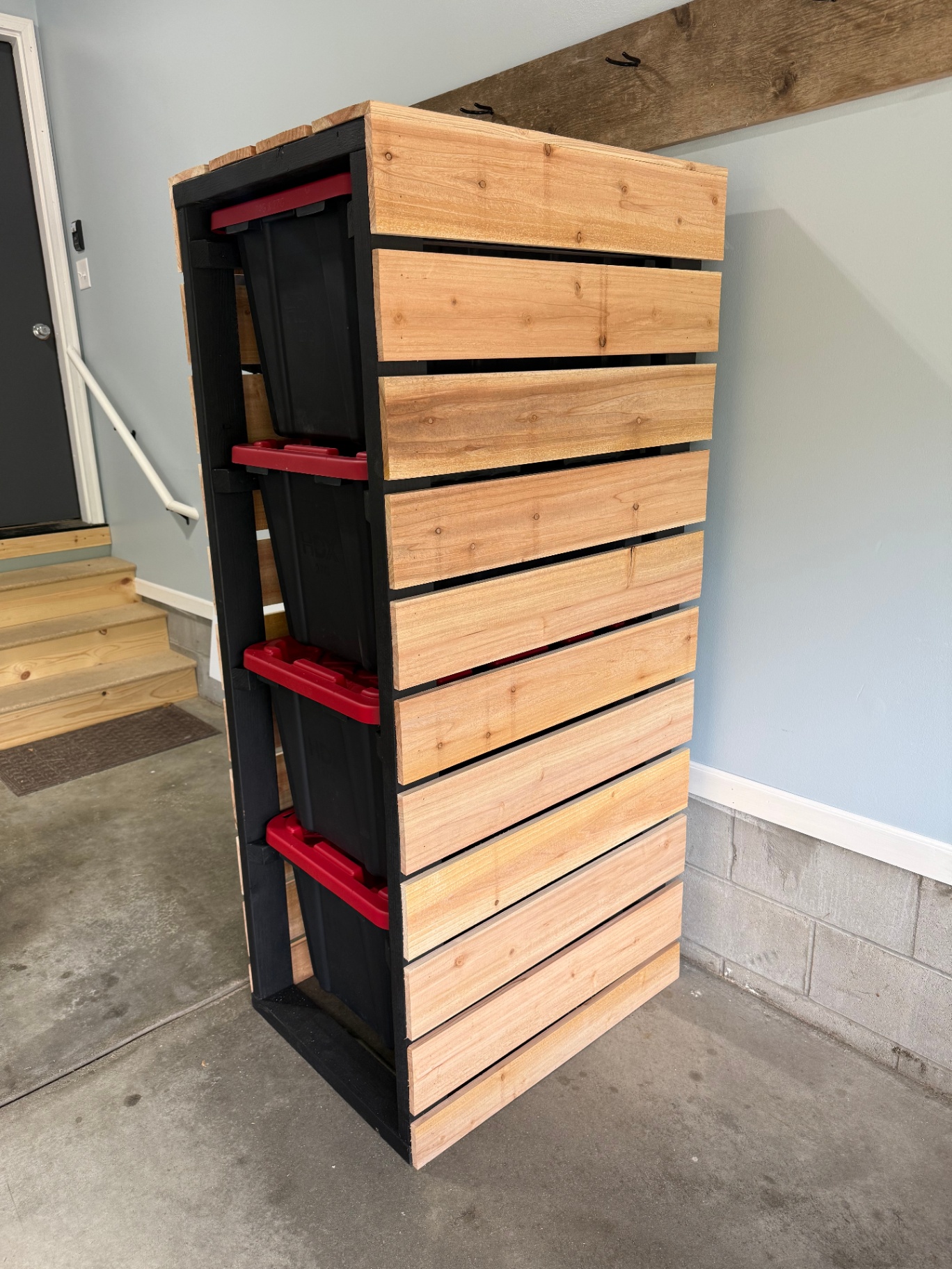 Tall custom-built garage storage rack with a black frame and horizontal cedar slat cladding holding stacked black storage totes